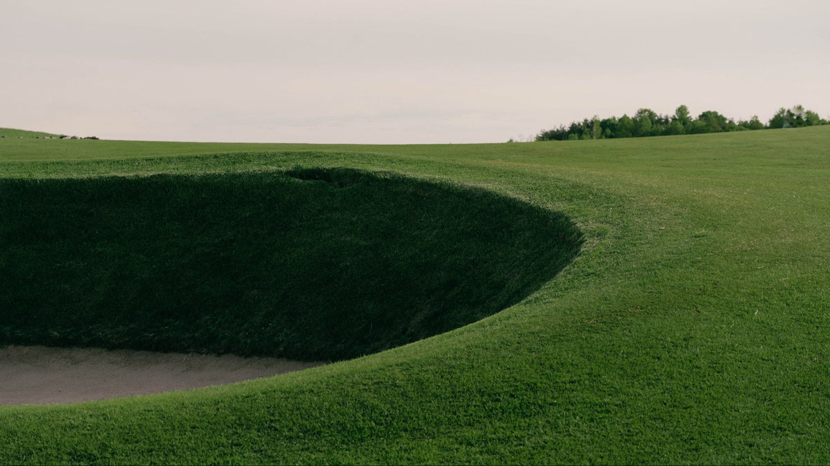 A scenic landscape photograph of a golf green hazard, featuring a deep, curved bunker or sand bunker on the left, a vibrant green grass fairway on the right, and a line of trees under a cloudy, overcast sky.